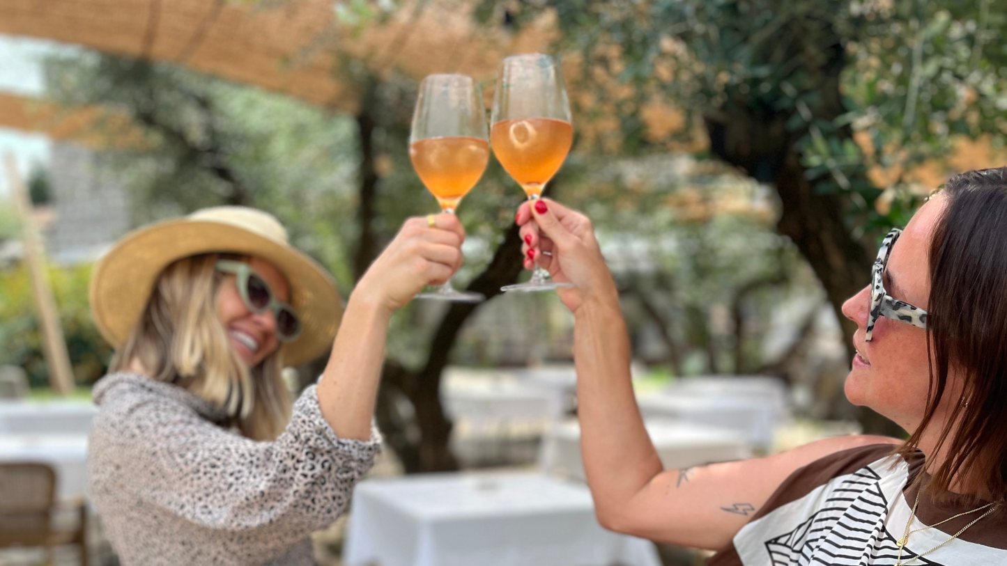 Two women in an outdoor patio setting clinking wine glasses filled with a light pink beverage. One woman wears a straw hat and sunglasses, and the other wears patterned sunglasses and a striped top.