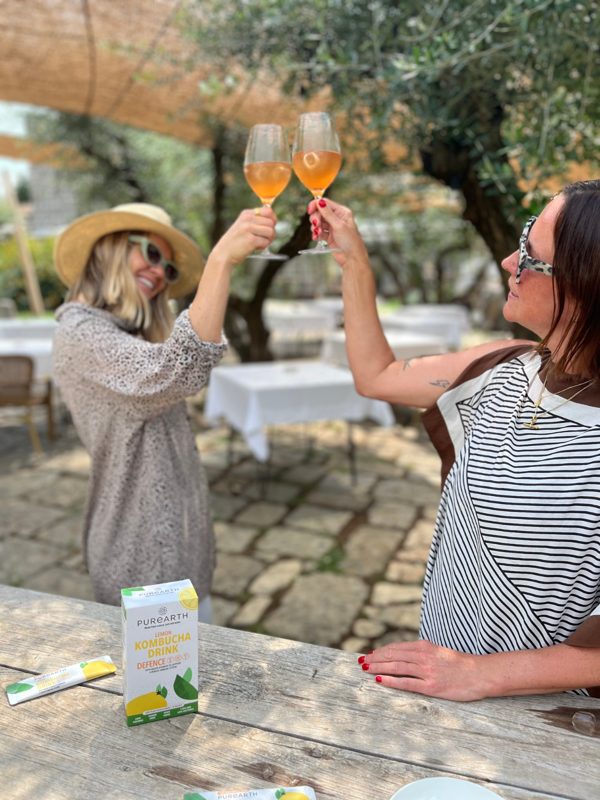 Two women clinking glasses of orange-colored beverage in an outdoor patio setting. A box of Purcellin Lemon & Mint tea and a tube of the same product are visible on a wooden table in the foreground.
